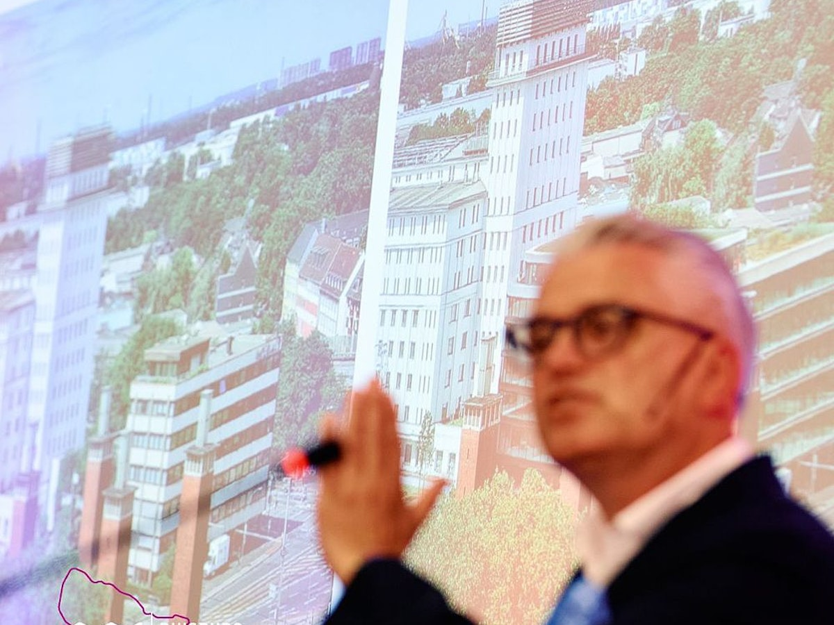 Eine Person gestikuliert während einer Präsentation vor einer Leinwand, auf der Stadtansichten und in der Ecke das Logo von „Duisburg Business Innovation“ zu sehen sind.