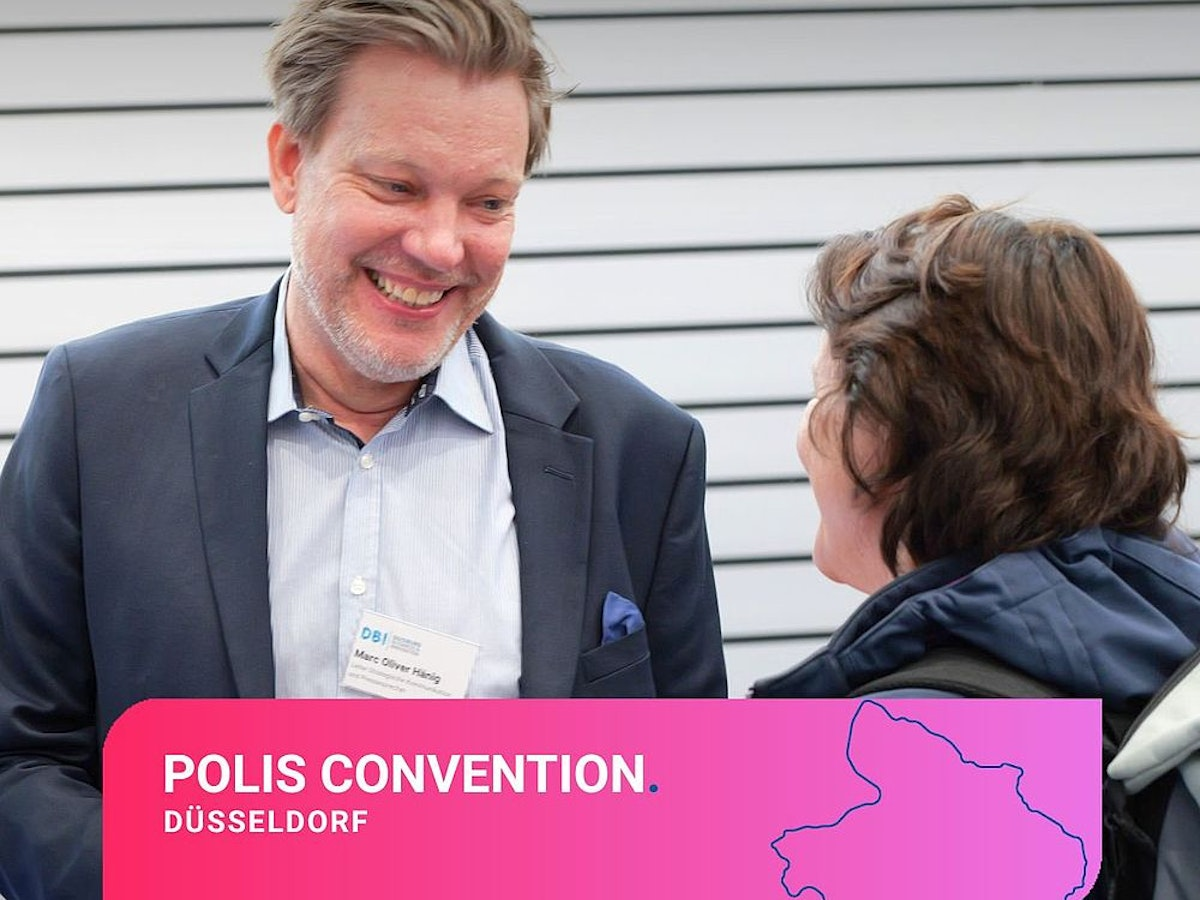 Auf der POLIS-Tagung in Düsseldorf stehen zwei Personen und lächeln einander an. Beide tragen Business-Kleidung und Namensschilder. Am unteren Bildrand ist ein rosa Banner mit Veranstaltungsdetails und einem Kartenausschnitt zu sehen.