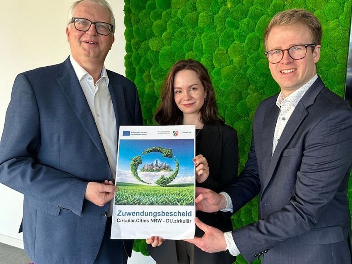 Drei Personen in Geschäftskleidung stehen lächelnd da und halten ein Zertifikat mit grünen Grafiken in den Händen. Sie posieren vor einer grünen Pflanzenwand. Das Logo von „Duisburg Business & Innovation“ befindet sich in der unteren linken Ecke.