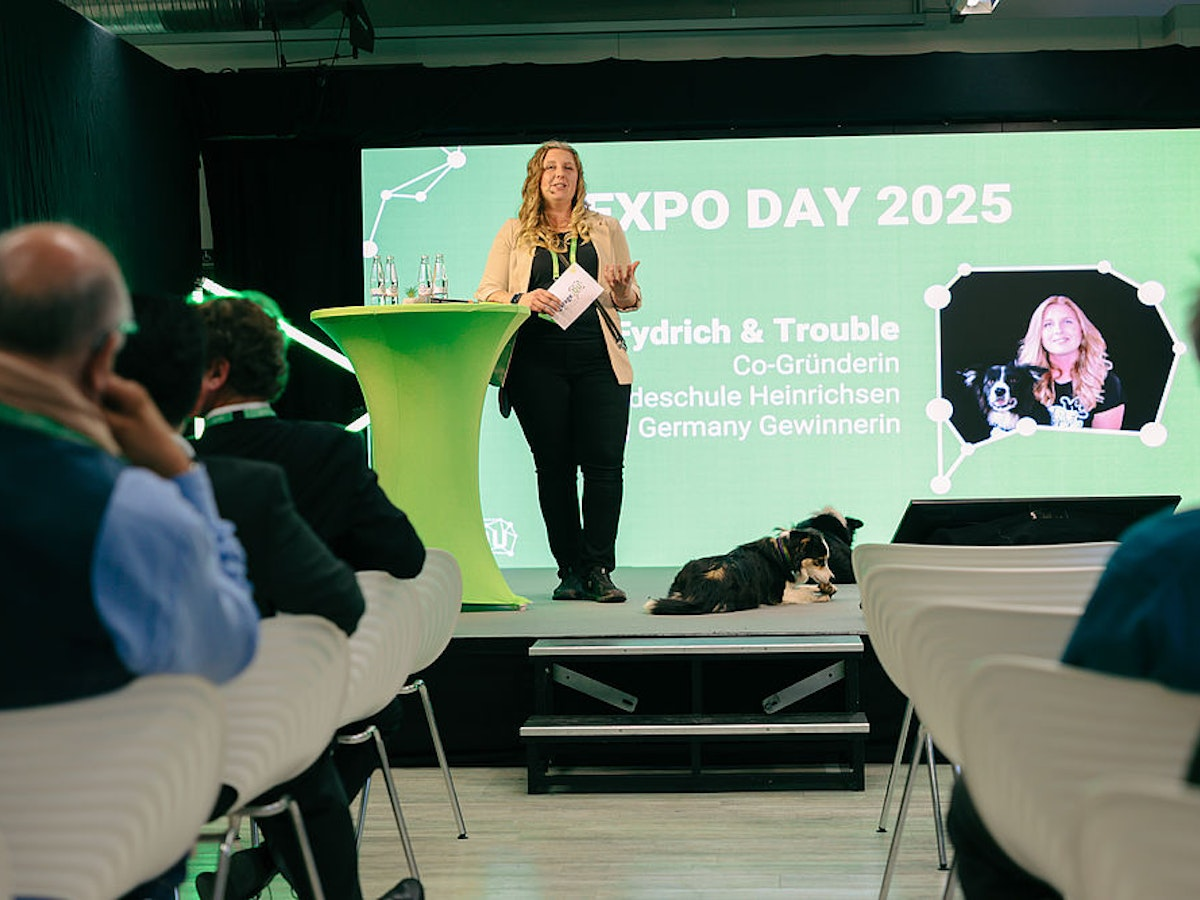 Eine Frau spricht auf der Bühne beim EXPO DAY 2025, neben ihr sitzt ein Hund, und das Publikum schaut ihr zu. Auf einem großen Bildschirm im Hintergrund sind ihr Foto mit dem Hund sowie Veranstaltungsdetails auf Deutsch zu sehen.