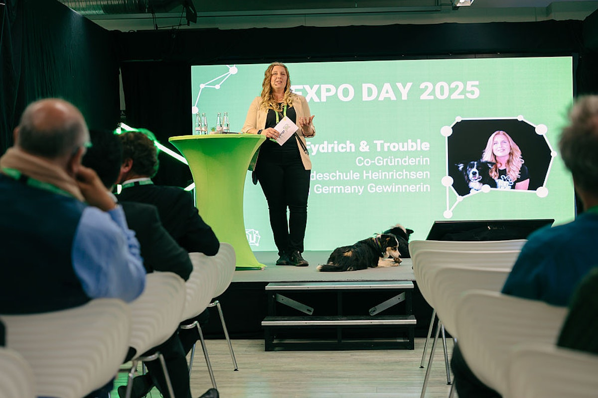 Eine Frau spricht auf der Bühne beim EXPO DAY 2025, neben ihr sitzt ein Hund, und das Publikum schaut ihr zu. Auf einem großen Bildschirm im Hintergrund sind ihr Foto mit dem Hund sowie Veranstaltungsdetails auf Deutsch zu sehen.