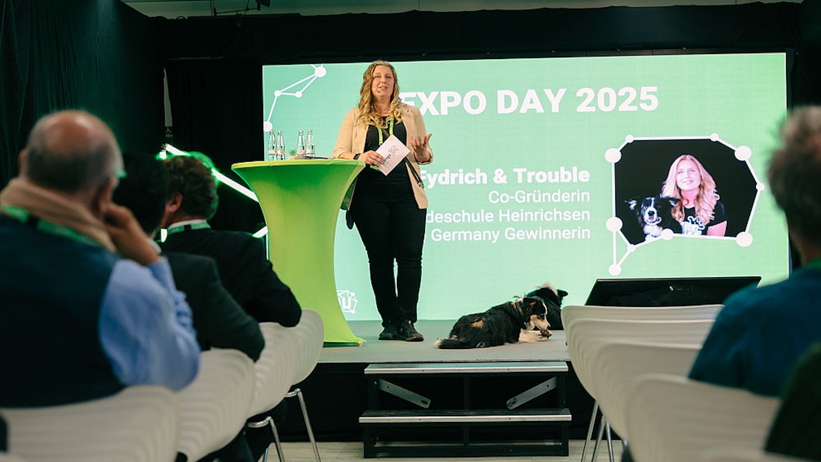 Eine Frau spricht auf der Bühne beim EXPO DAY 2025, neben ihr sitzt ein Hund, und das Publikum schaut ihr zu. Auf einem großen Bildschirm im Hintergrund sind ihr Foto mit dem Hund sowie Veranstaltungsdetails auf Deutsch zu sehen.