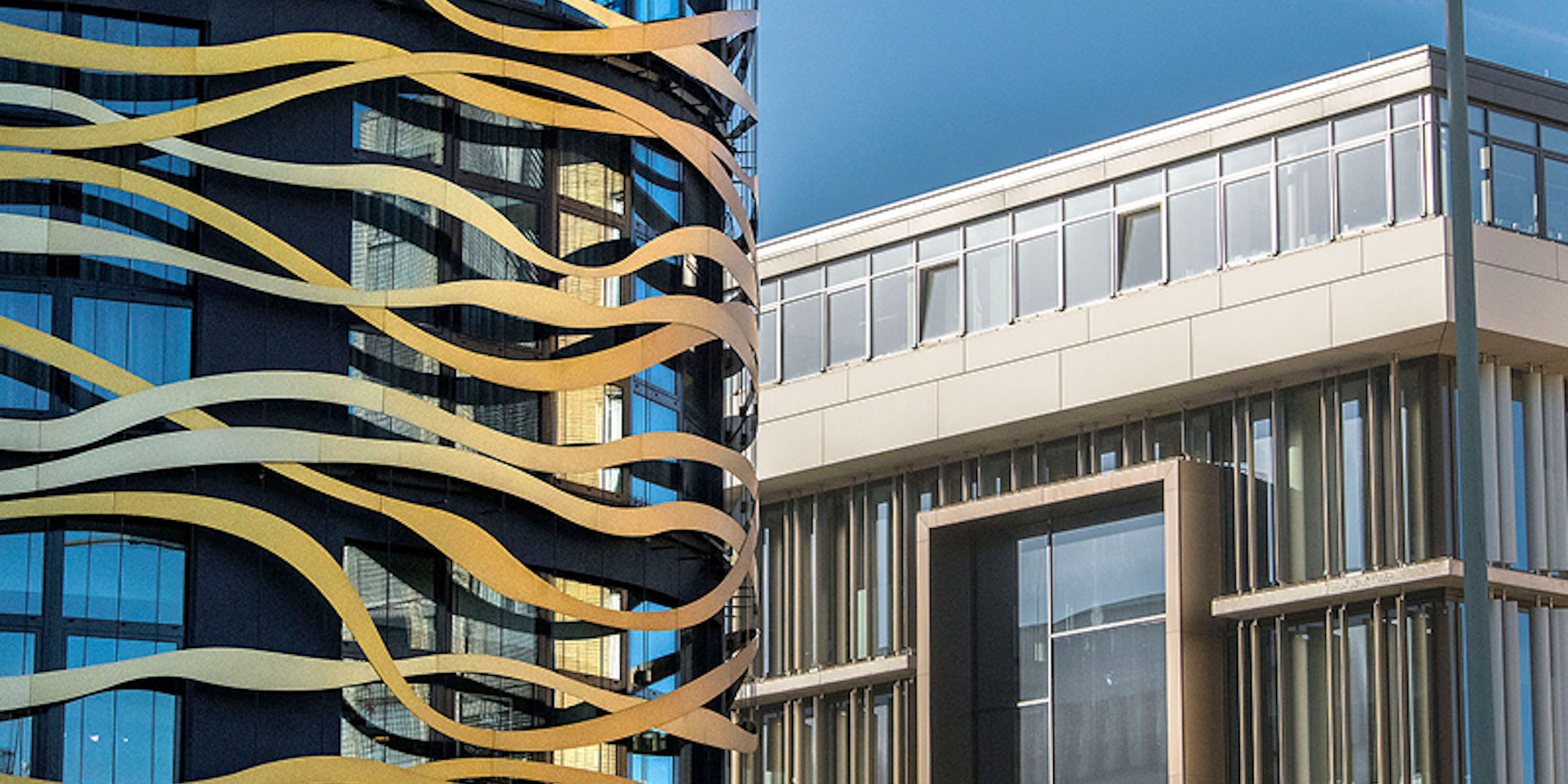 Ein modernes schwarzes Gebäude mit goldenen Wellenlinien steht neben einem beigen, rechteckigen Gebäude mit großen Fenstern und einem markanten, kastenförmigen Fensterelement. Im Hintergrund ist der blaue Himmel zu sehen.