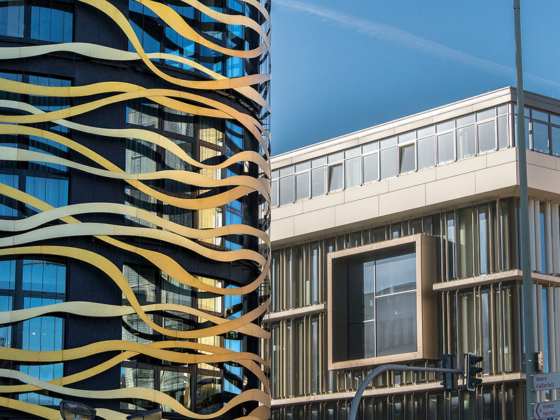 Ein modernes schwarzes Gebäude mit goldenen Wellenlinien steht neben einem beigen, rechteckigen Gebäude mit großen Fenstern und einem markanten, kastenförmigen Fensterelement. Im Hintergrund ist der blaue Himmel zu sehen.