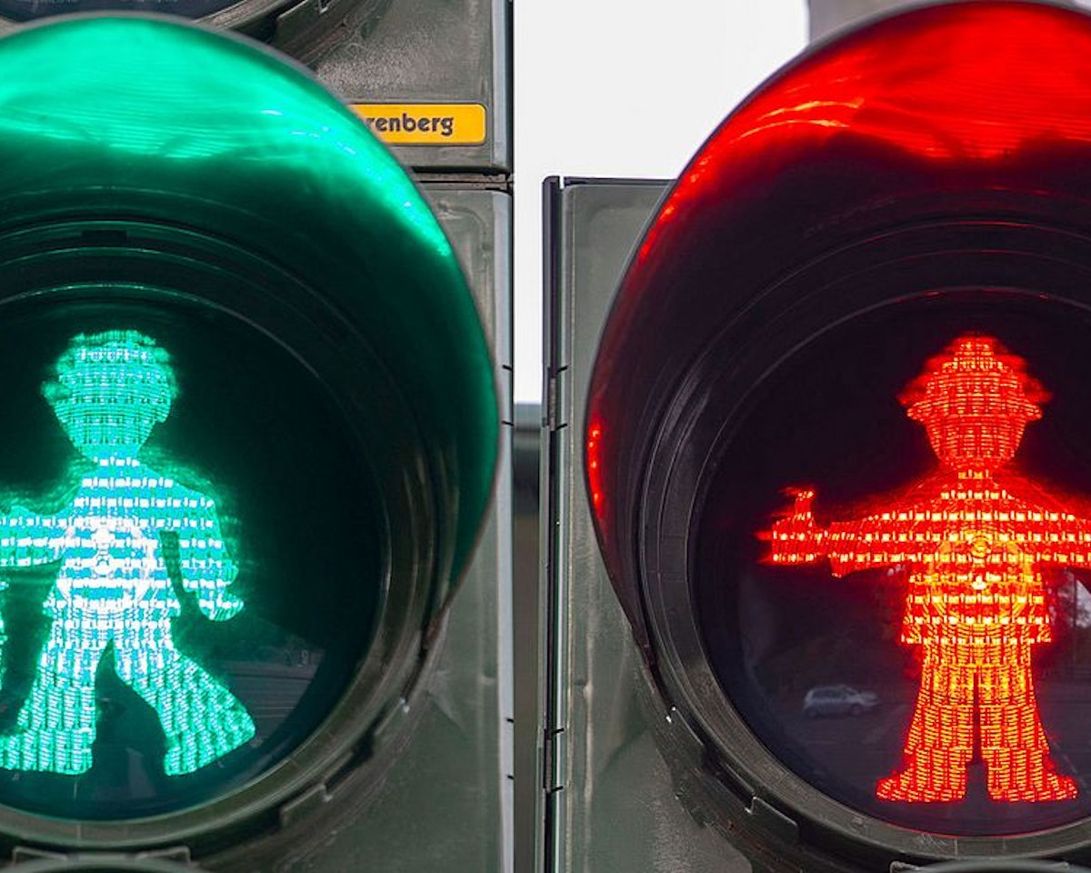Ein geteiltes Bild, das grüne und rote Fußgängerampeln mit einzigartigen Symbolen zeigt: Das grüne Symbol zeigt eine gehende Person, das rote eine stehende Person, die in beiden Händen Gegenstände hält.