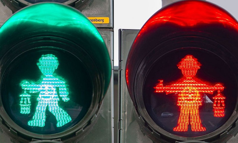 Ein geteiltes Bild, das grüne und rote Fußgängerampeln mit einzigartigen Symbolen zeigt: Das grüne Symbol zeigt eine gehende Person, das rote eine stehende Person, die in beiden Händen Gegenstände hält.