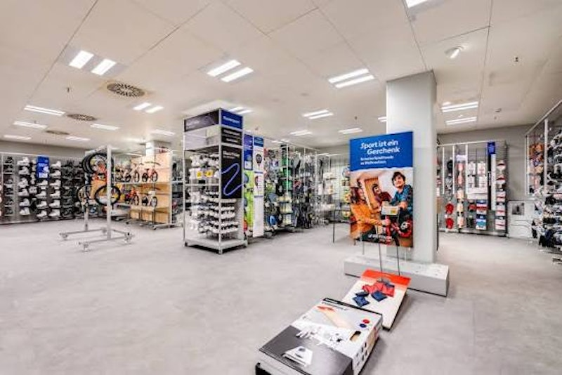 Ein hell beleuchtetes Sportgeschäft mit Regalen und Ständern, in denen verschiedene Sportgeräte und Accessoires ausgestellt sind. Im gesamten geräumigen, übersichtlichen Laden sind Schilder und Plakate zu sehen.