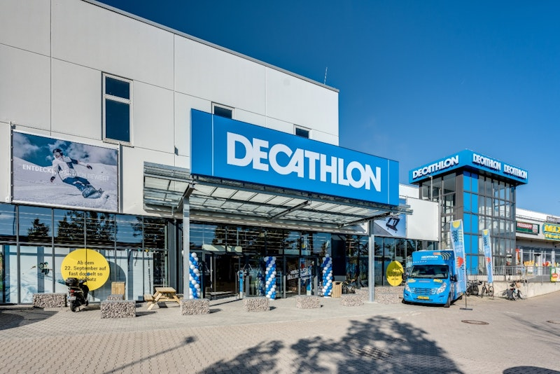 Außenansicht einer Decathlon-Filiale mit großem blauem Schild, gläsernem Eingangsbereich, einem geparkten blauen Fahrzeug und Werbebannern; modernes Gebäude unter klarem Himmel.