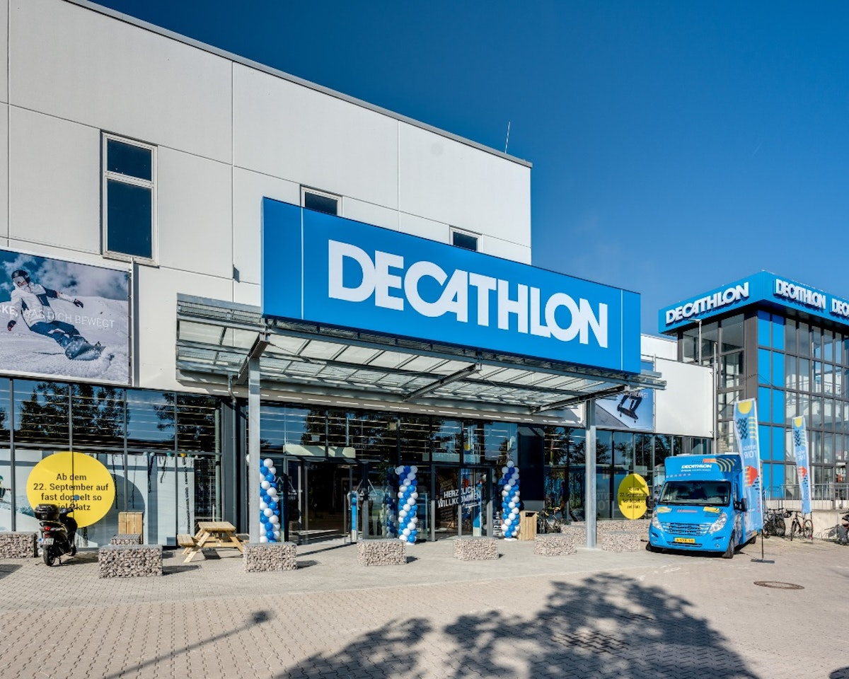 Außenansicht einer Decathlon-Filiale mit großem blauem Schild, gläsernem Eingangsbereich, einem geparkten blauen Fahrzeug und Werbebannern; modernes Gebäude unter klarem Himmel.