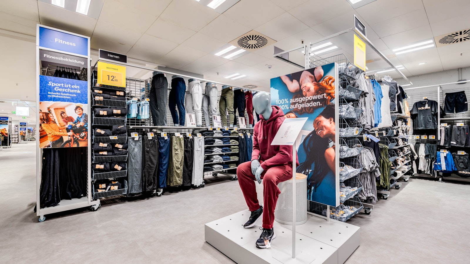 Eine Schaufensterpuppe in einem roten Trainingsanzug sitzt auf einer Ausstellungsfläche in einem hell beleuchteten Bekleidungsgeschäft, umgeben von Regalen mit Hosen und Fitnessbekleidung. Im gesamten Geschäft sind Werbeschilder und gefaltete Kleidungsstücke ordentlich angeordnet.
