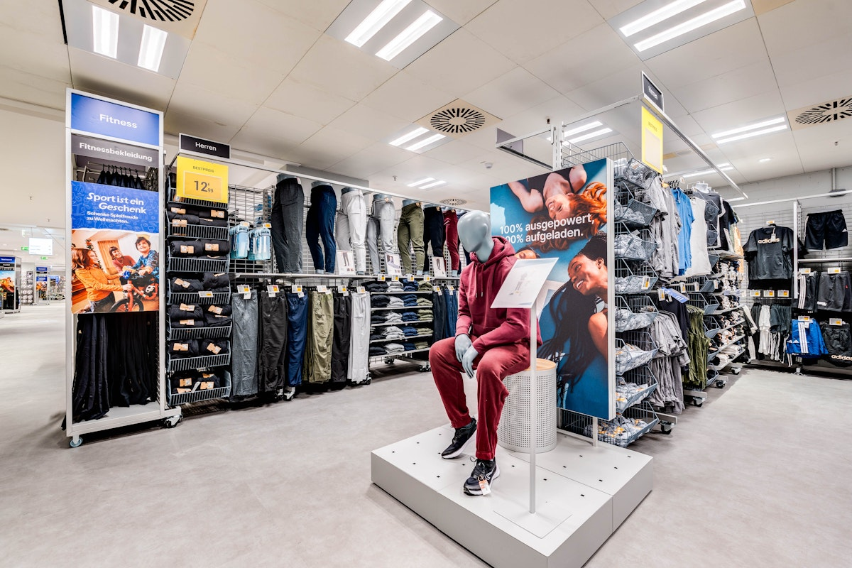 Eine Schaufensterpuppe in einem roten Trainingsanzug sitzt auf einer Ausstellungsfläche in einem hell beleuchteten Bekleidungsgeschäft, umgeben von Regalen mit Hosen und Fitnessbekleidung. Im gesamten Geschäft sind Werbeschilder und gefaltete Kleidungsstücke ordentlich angeordnet.