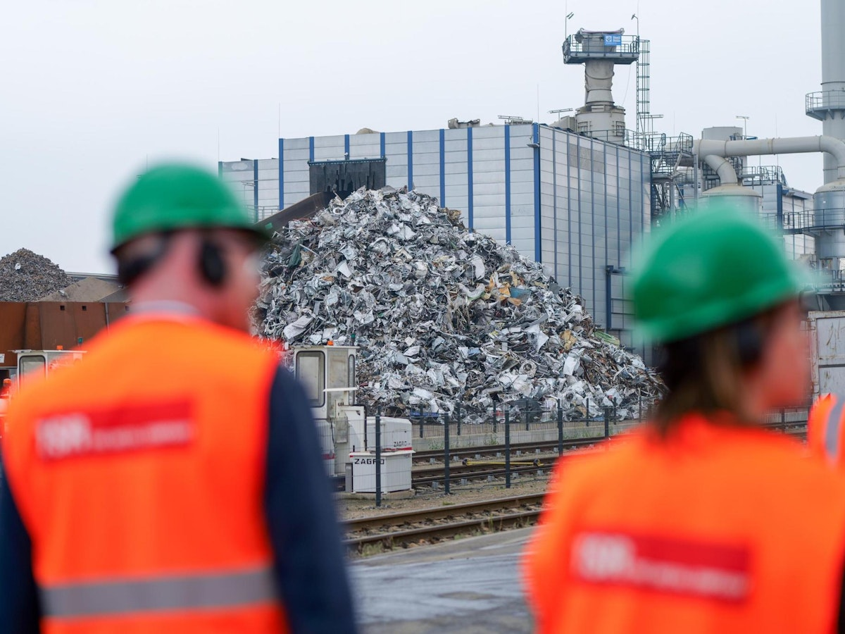 Arbeiter in orangefarbenen Westen und grünen Helmen stehen vor einem großen Haufen Metallschrott vor einer Industrieanlage, im Hintergrund sind Rohrleitungen und ein modernes Gebäude zu sehen.