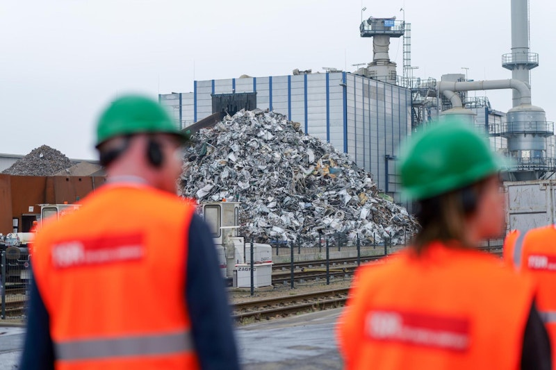 Arbeiter in orangefarbenen Westen und grünen Helmen stehen vor einem großen Haufen Metallschrott vor einer Industrieanlage, im Hintergrund sind Rohrleitungen und ein modernes Gebäude zu sehen.