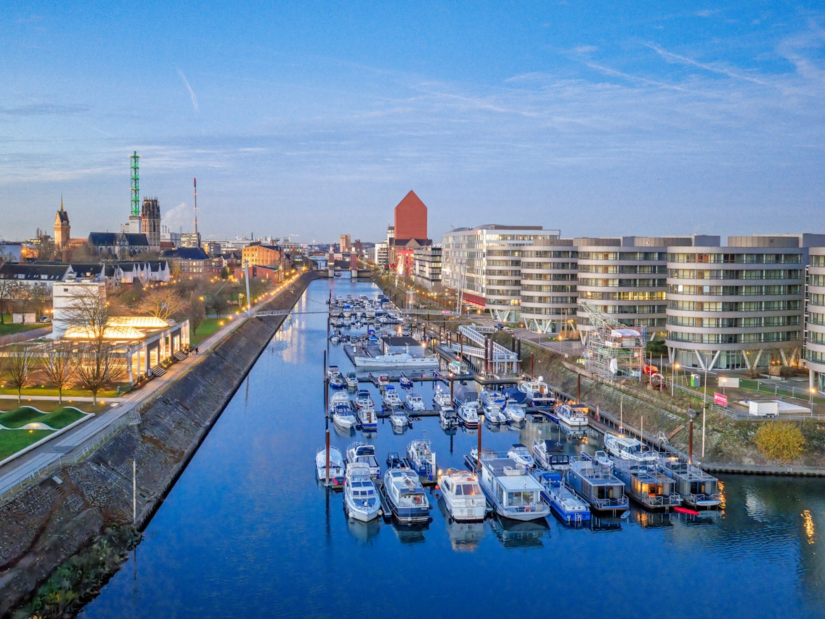 Luftaufnahme eines Yachthafens mit zahlreichen vor Anker liegenden Booten, umgeben von modernen Gebäuden und Grünflächen an einem ruhigen Fluss unter strahlend blauem Himmel.