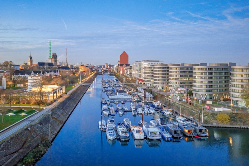 Luftaufnahme eines Yachthafens mit zahlreichen vor Anker liegenden Booten, umgeben von modernen Gebäuden und Grünflächen an einem ruhigen Fluss unter strahlend blauem Himmel.