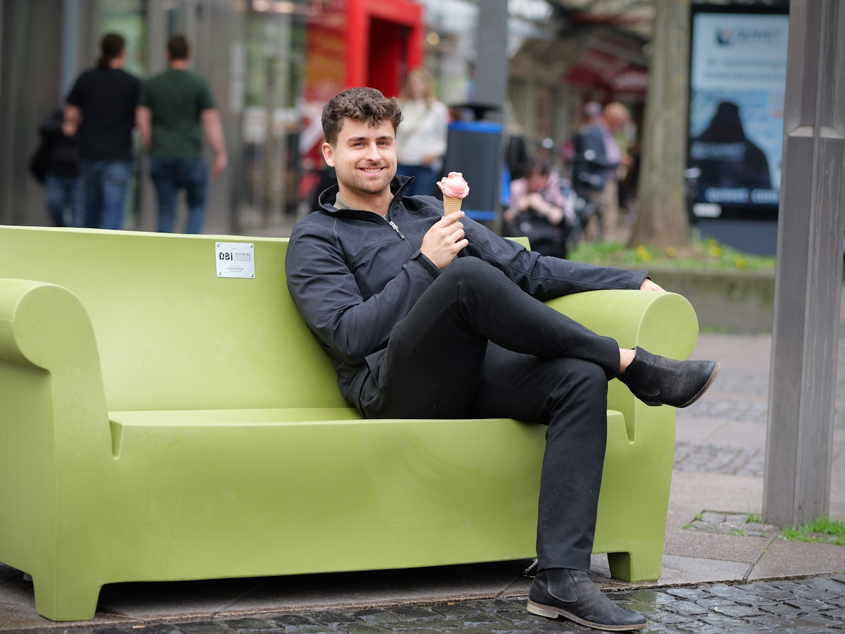 Ein Mann in schwarzer Kleidung sitzt auf einer großen grünen Bank, die wie ein Sofa geformt ist. Er lächelt und hält eine Eistüte in der Hand, während im Hintergrund Menschen in einer städtischen Außenanlage zu sehen sind.