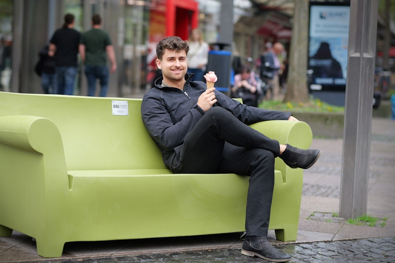 Ein Mann in schwarzer Kleidung sitzt auf einer großen grünen Bank, die wie ein Sofa geformt ist. Er lächelt und hält eine Eistüte in der Hand, während im Hintergrund Menschen in einer städtischen Außenanlage zu sehen sind.