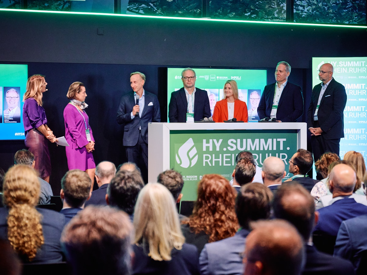 Sechs Personen in Geschäftskleidung sprechen auf einer Bühne mit der Aufschrift „HY.SUMMIT. RHEIN.RUHR“ vor einem Publikum, hinter ihnen sind eine Leinwand und Banner mit dem Namen der Veranstaltung zu sehen.
