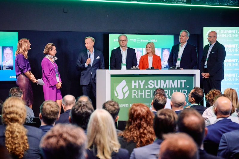 Sechs Personen in Geschäftskleidung sprechen auf einer Bühne mit der Aufschrift „HY.SUMMIT. RHEIN.RUHR“ vor einem Publikum, hinter ihnen sind eine Leinwand und Banner mit dem Namen der Veranstaltung zu sehen.