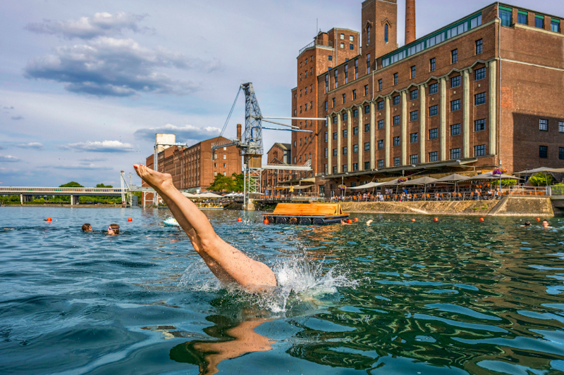 Eine Person springt in der Nähe eines großen Backsteingebäudes in einen Fluss und verursacht dabei einen Spritzer. Andere Menschen schwimmen in der Nähe, und Zuschauer sitzen in einem Straßencafé am Ufer.