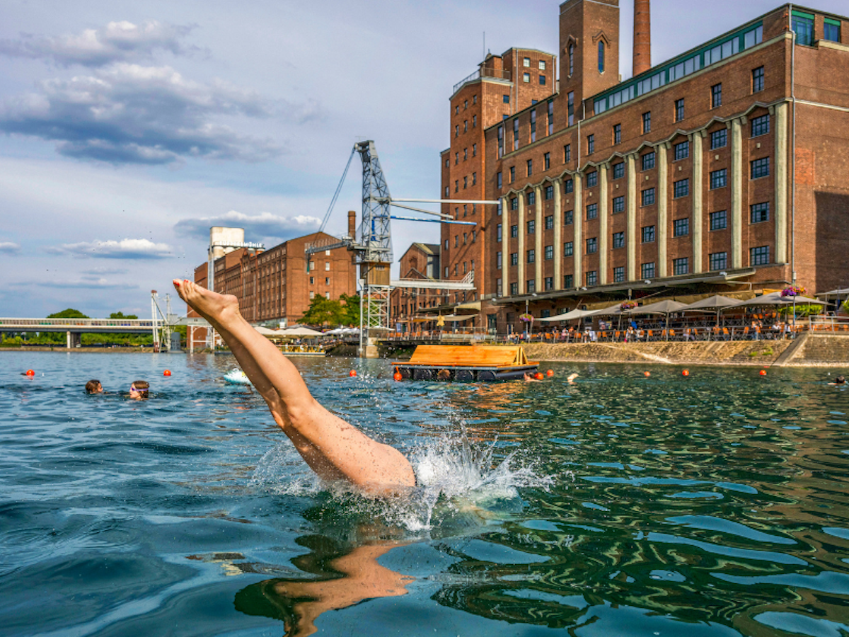 Eine Person springt in der Nähe eines großen Backsteingebäudes in einen Fluss und verursacht dabei einen Spritzer. Andere Menschen schwimmen in der Nähe, und Zuschauer sitzen in einem Straßencafé am Ufer.