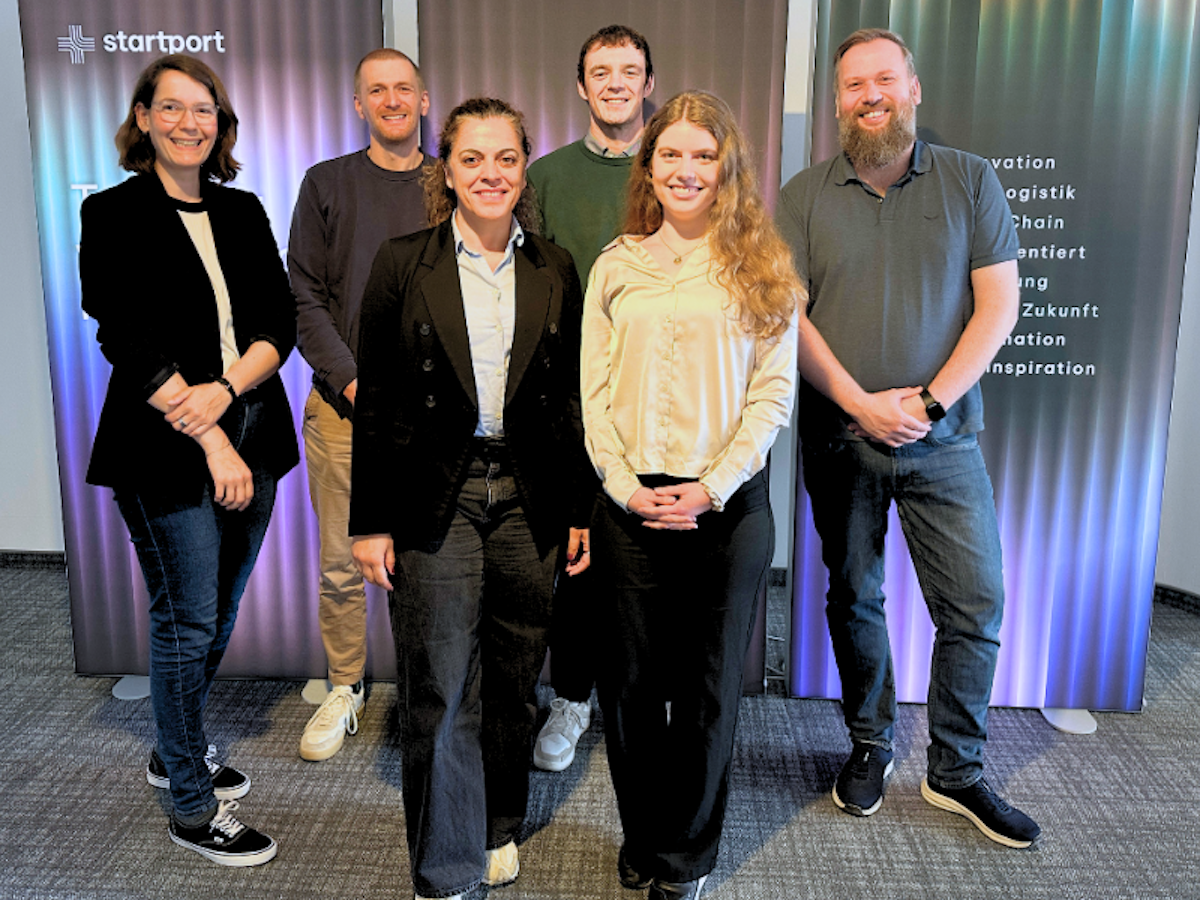 Sechs Personen stehen in Business-Casual-Kleidung vor einem violett und grün beleuchteten Hintergrund, auf dem das Wort „startport“ und ein Text auf Deutsch zu sehen sind, und lächeln in die Kamera.