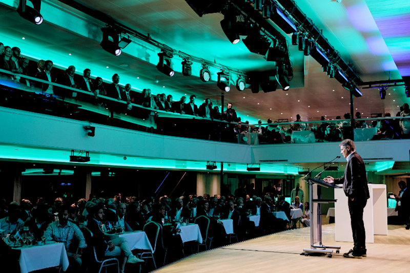 Ein Redner steht am Rednerpult und spricht zu einem großen Publikum, das an runden Tischen in einem modernen, gut beleuchteten Konferenzsaal mit zwei Ebenen sitzt.