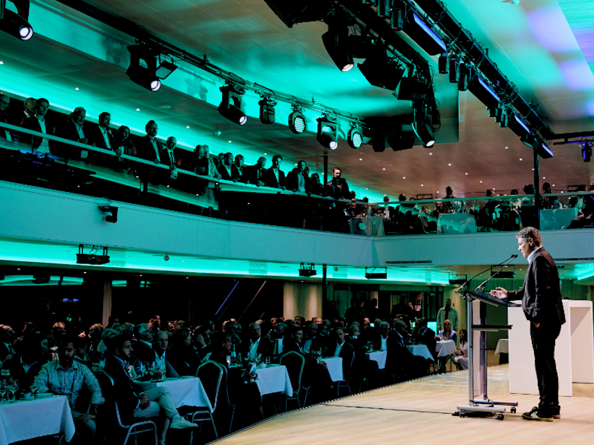 Ein Redner steht am Rednerpult und spricht zu einem großen Publikum, das an runden Tischen in einem modernen, gut beleuchteten Konferenzsaal mit zwei Ebenen sitzt.