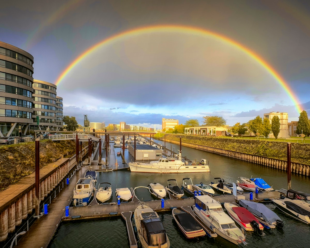 Unterhalb eines leuchtenden Doppelregenbogens, der sich über moderne Gebäude und einen bewölkten Himmel spannt, liegt ein Yachthafen mit vor Anker liegenden Booten, während Sonnenlicht die Szene erhellt.