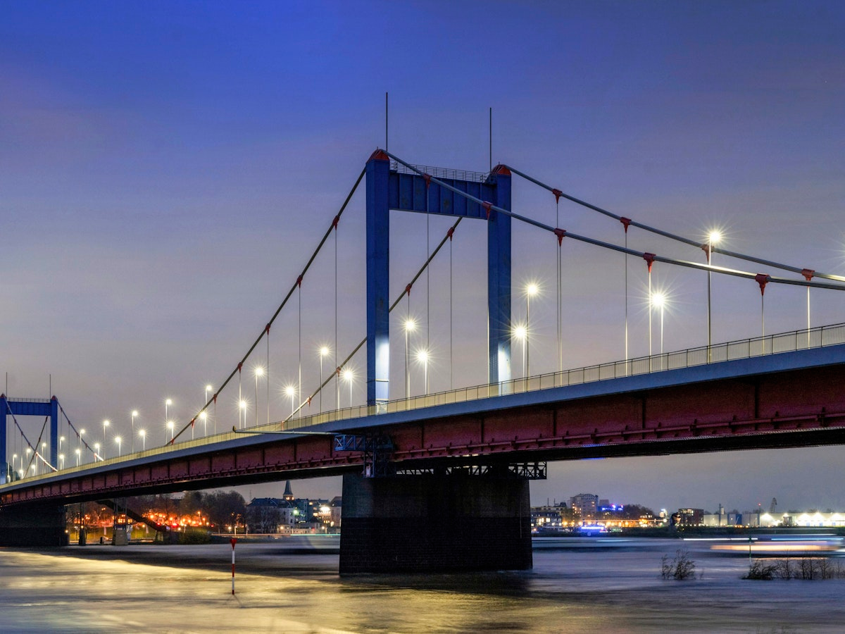 Eine von Straßenlaternen beleuchtete Hängebrücke überspannt in der Abenddämmerung einen breiten Fluss, im Hintergrund sind die Skyline der Stadt und Spiegelungen zu sehen.