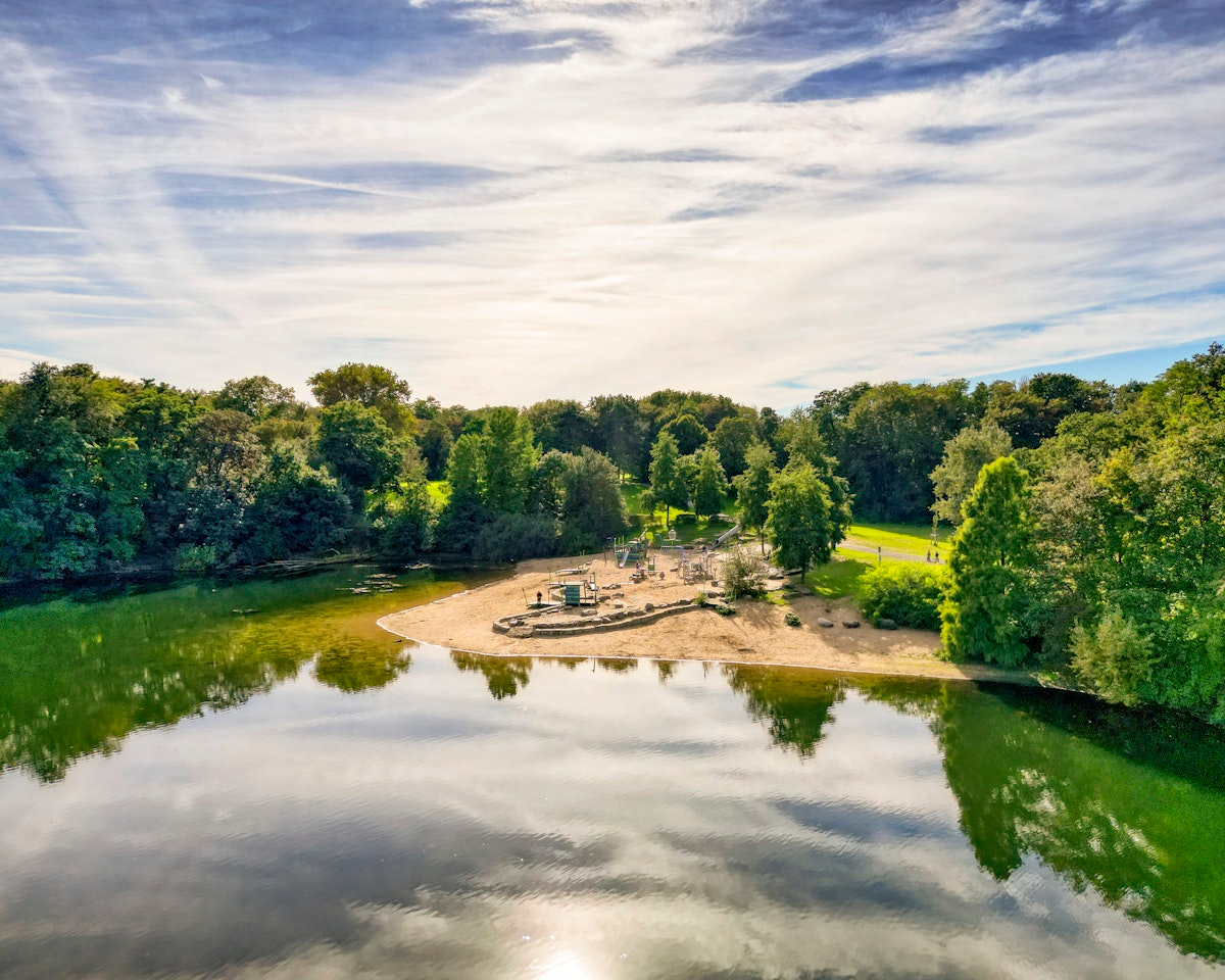 Ein Sandstrand liegt an einem ruhigen, spiegelglatten See, umgeben von üppig grünen Bäumen unter einem teilweise bewölkten Himmel; man sieht Menschen, die die Natur genießen.
