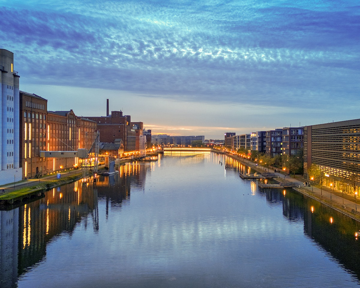 In der Abenddämmerung fließt ein Fluss durch eine Stadt und spiegelt die Lichter der Gebäude auf beiden Seiten wider. Der Himmel ist blau und mit vereinzelten Wolken überzogen, und in der Ferne ist eine Brücke zu sehen. Auf dem Gebäude links steht „KÜPPERSMÜHLE“.
