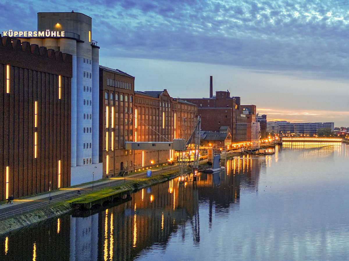 Ein Blick vom Ufer auf das Gebäude des Museums Küppersmühle in der Abenddämmerung, mit Lichtern, die sich auf dem ruhigen Fluss spiegeln, und einem teilweise bewölkten Himmel darüber.