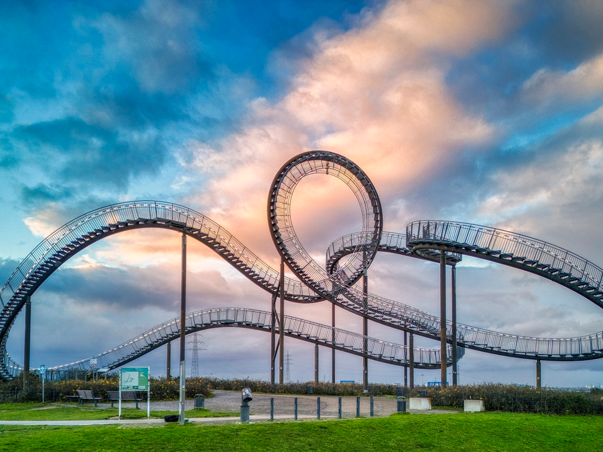 Eine sich windende, geschwungene Metallkonstruktion, die als Fußgängerbrücke dient, steht auf grünem Gras unter einem dramatischen Wolkenhimmel. Eine Person beobachtet das Geschehen aus der Ferne.