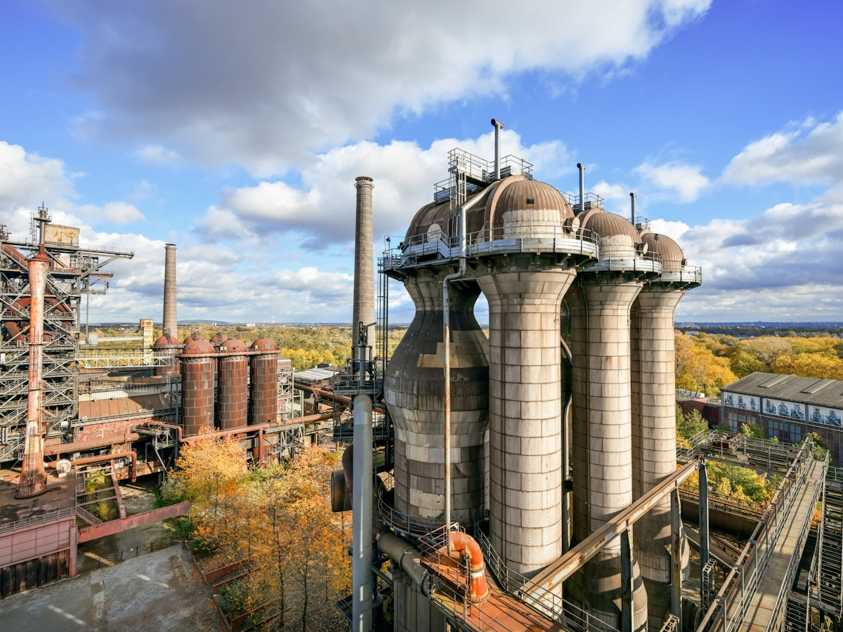 Ein großes Stahlwerk mit hohen zylindrischen Türmen und Rohren, umgeben von herbstlichen Bäumen unter einem teilweise bewölkten Himmel. Industriegebäude und -anlagen prägen die Landschaft.
