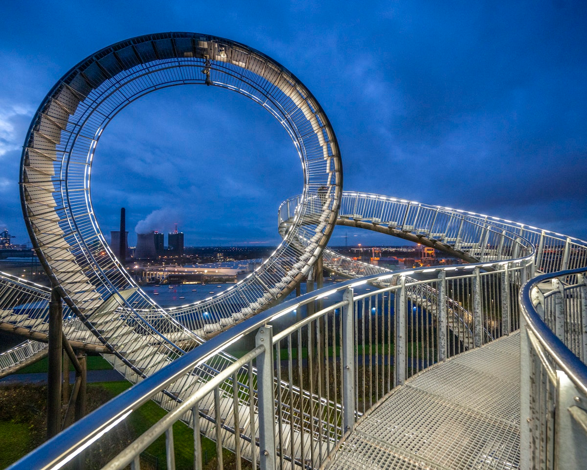 Ein großer, geschwungener Metallsteg, der einer Achterbahn ähnelt, schlängelt sich in der Abenddämmerung dramatisch vor dem Hintergrund der Stadtlandschaft entlang und wird von Lichtern entlang seiner Geländer beleuchtet.