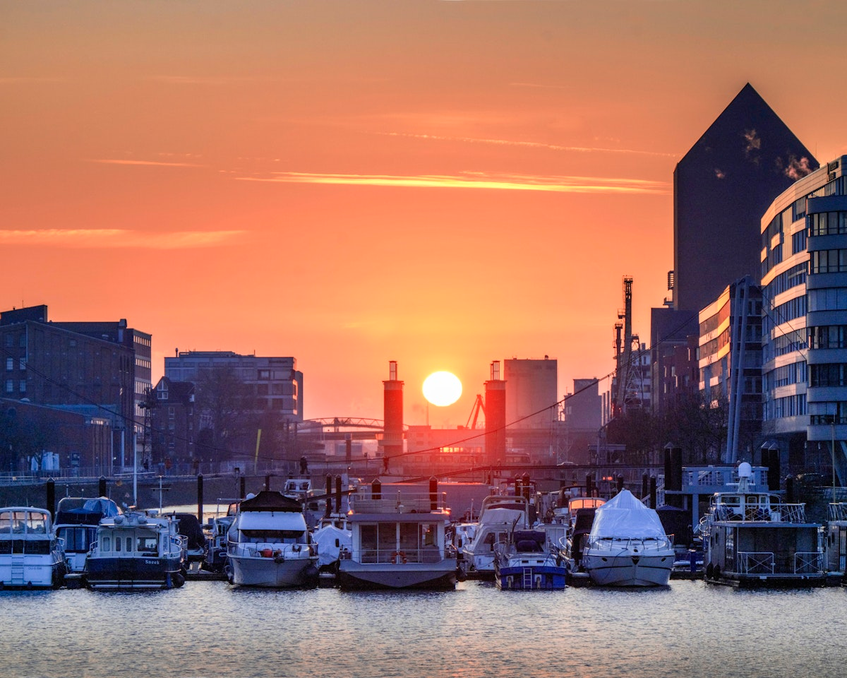 Bei Sonnenuntergang lagen Boote im Hafen vor Anker, während die Sonne zwischen modernen und älteren Gebäuden unterging und einen orangefarbenen Schimmer über das Wasser und den Himmel warf.