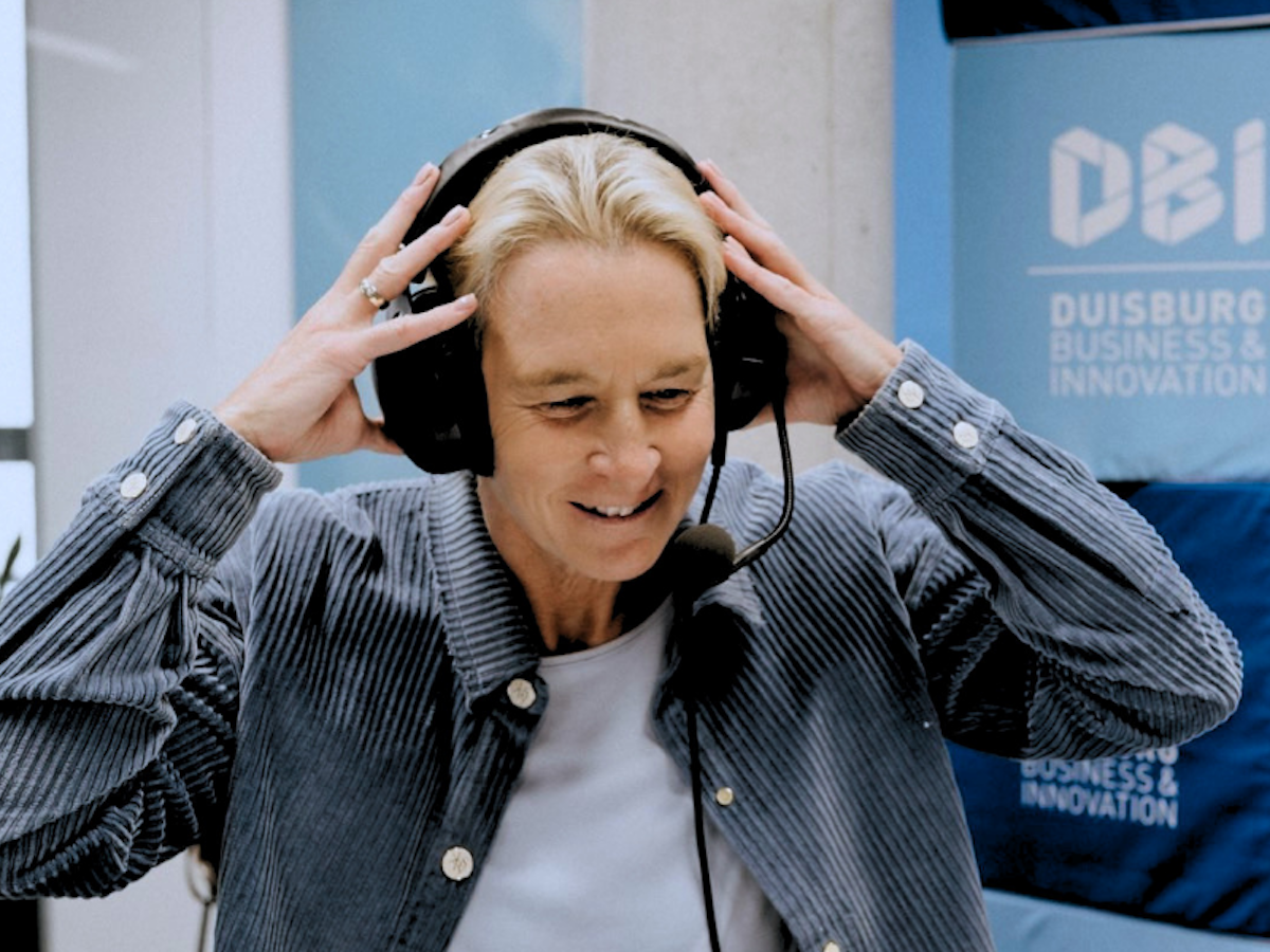 Eine Person, die ein Headset und ein Mikrofon trägt, lächelt, während sie das Headset zurechtzieht. Hinter ihr ist auf einem blauen Banner die Aufschrift „Duisburg Business & Innovation“ zu sehen. Die Person scheint gerade an einer Sendung oder einem Interview teilzunehmen.