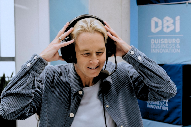 Eine Person, die ein Headset und ein Mikrofon trägt, lächelt, während sie das Headset zurechtzieht. Hinter ihr ist auf einem blauen Banner die Aufschrift „Duisburg Business & Innovation“ zu sehen. Die Person scheint gerade an einer Sendung oder einem Interview teilzunehmen.